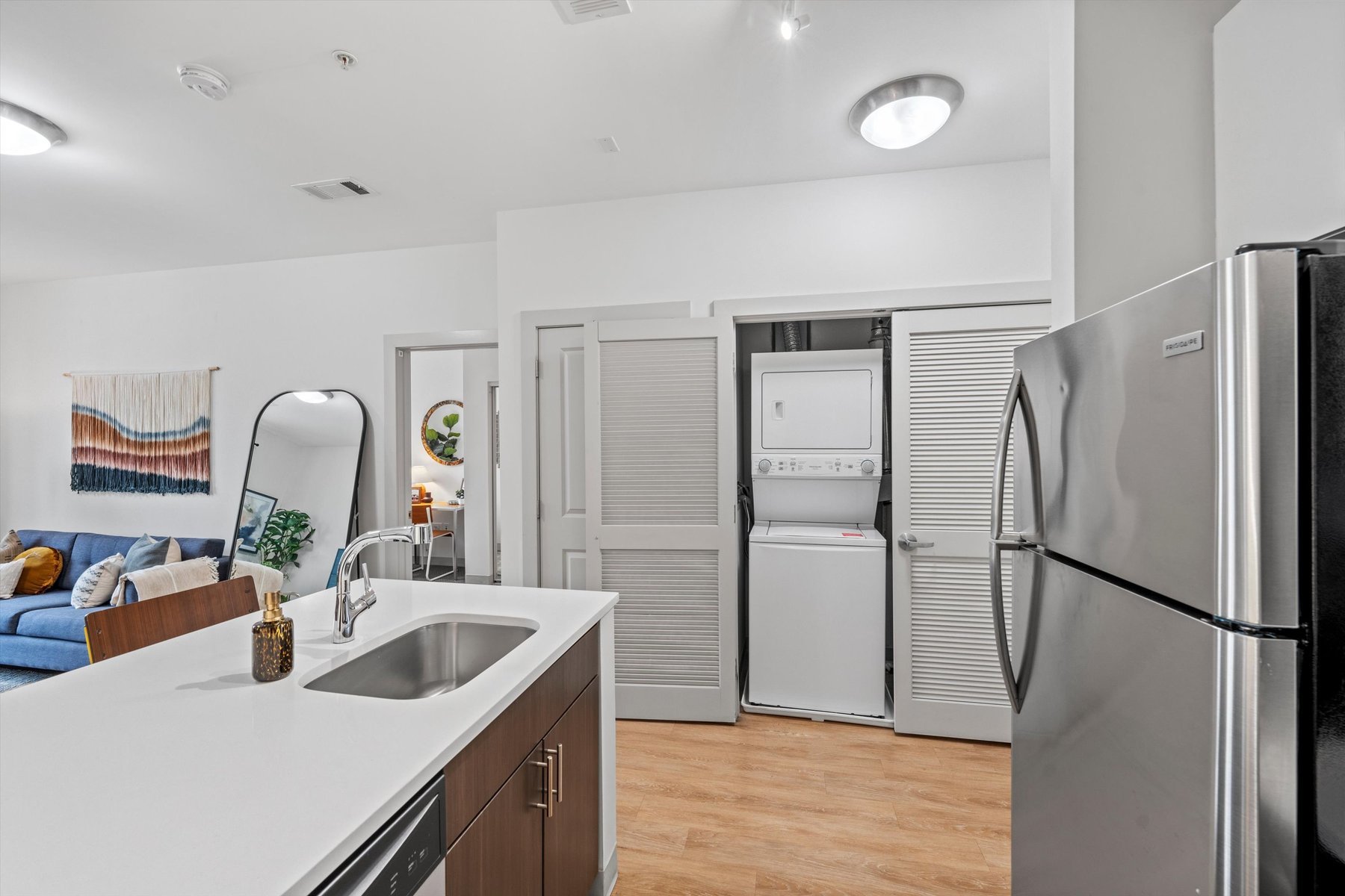 Modern apartment kitchen with stainless steel appliances and a sink island at Landmark College Park; in the background, an open closet reveals a stacked washer and dryer, plus a living area with a blue sofa and wall art.