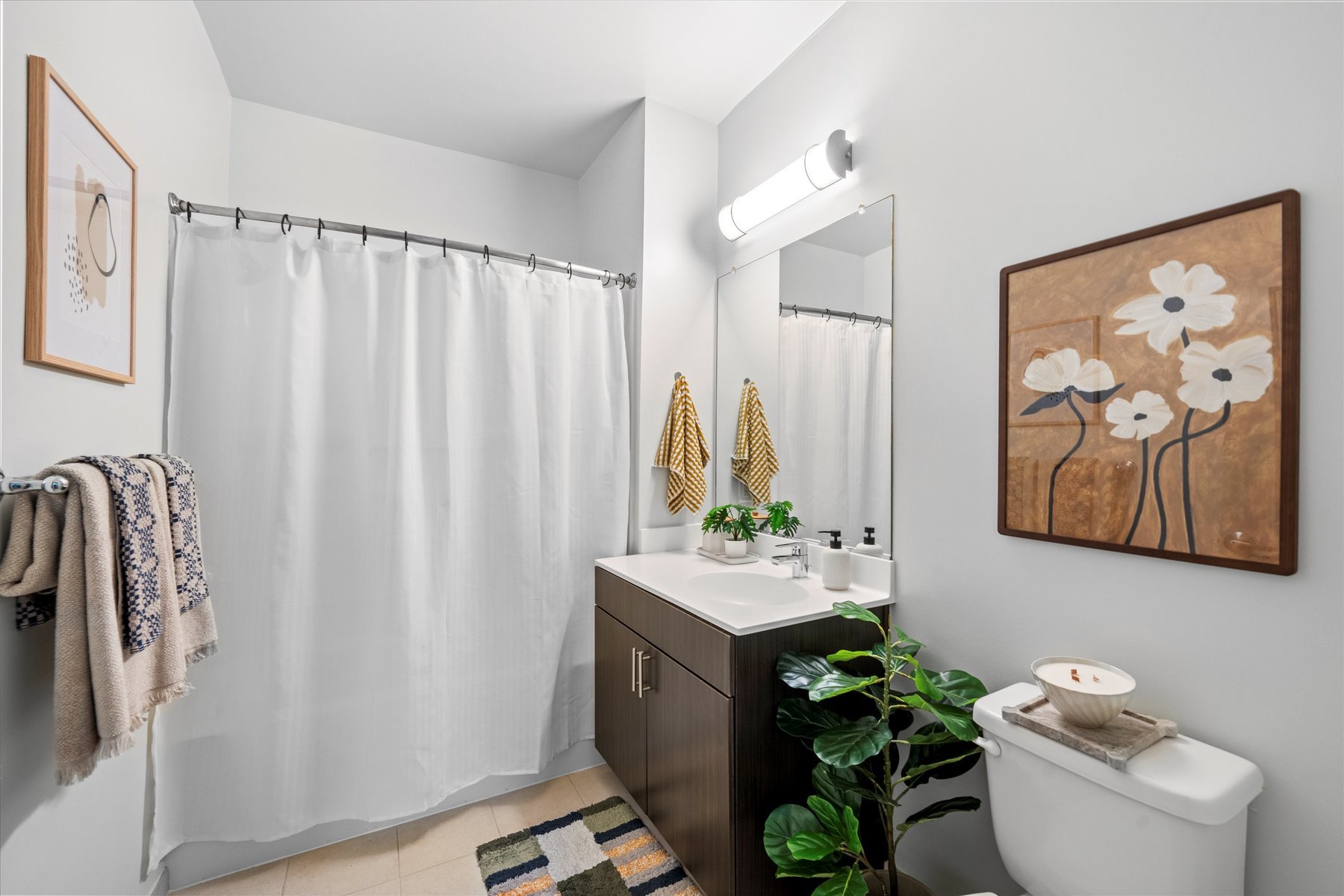 Modern bathroom at Landmark College Park features a white shower curtain, dark vanity with sink, large mirror, towel rack, toilet, potted plant, and flower-themed wall art. Towels and a striped rug add color to the neutral space.