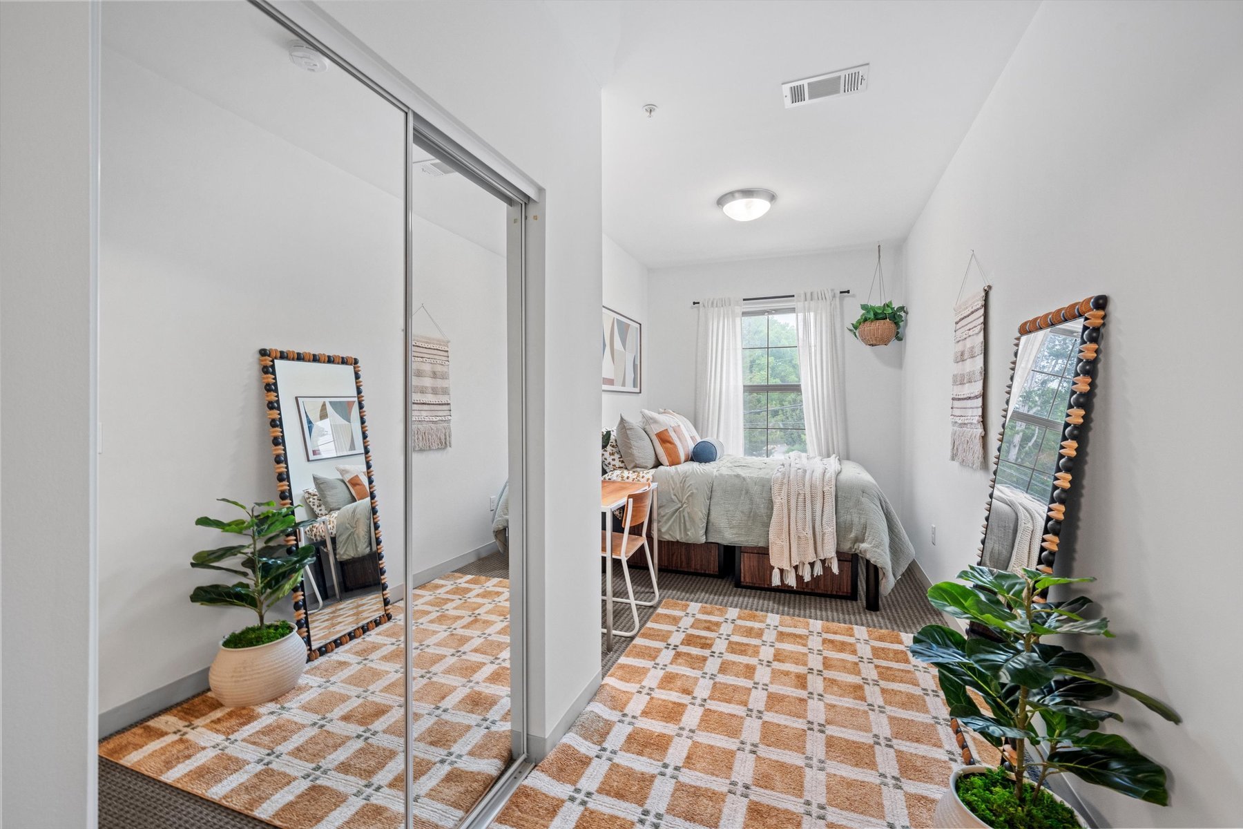 A bright, modern bedroom at Landmark College Park features a bed, desk, chair, potted plants, large mirror, and a window with sheer curtains. Earthy tones and a checkered rug complete the look, with mirrored closet doors on the left.