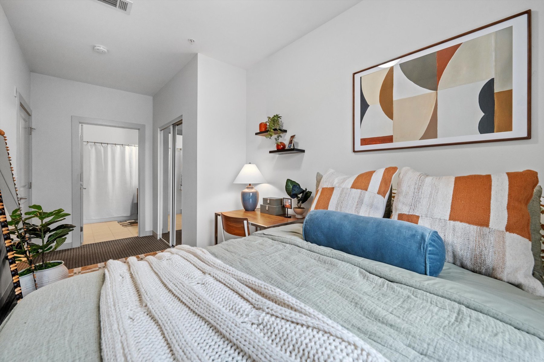 A cozy bedroom at Landmark College Park features a neatly made bed with striped pillows and a blue bolster. A desk with a lamp and plants sits against the wall, modern art hangs above the bed, and a bathroom is visible through an open doorway.