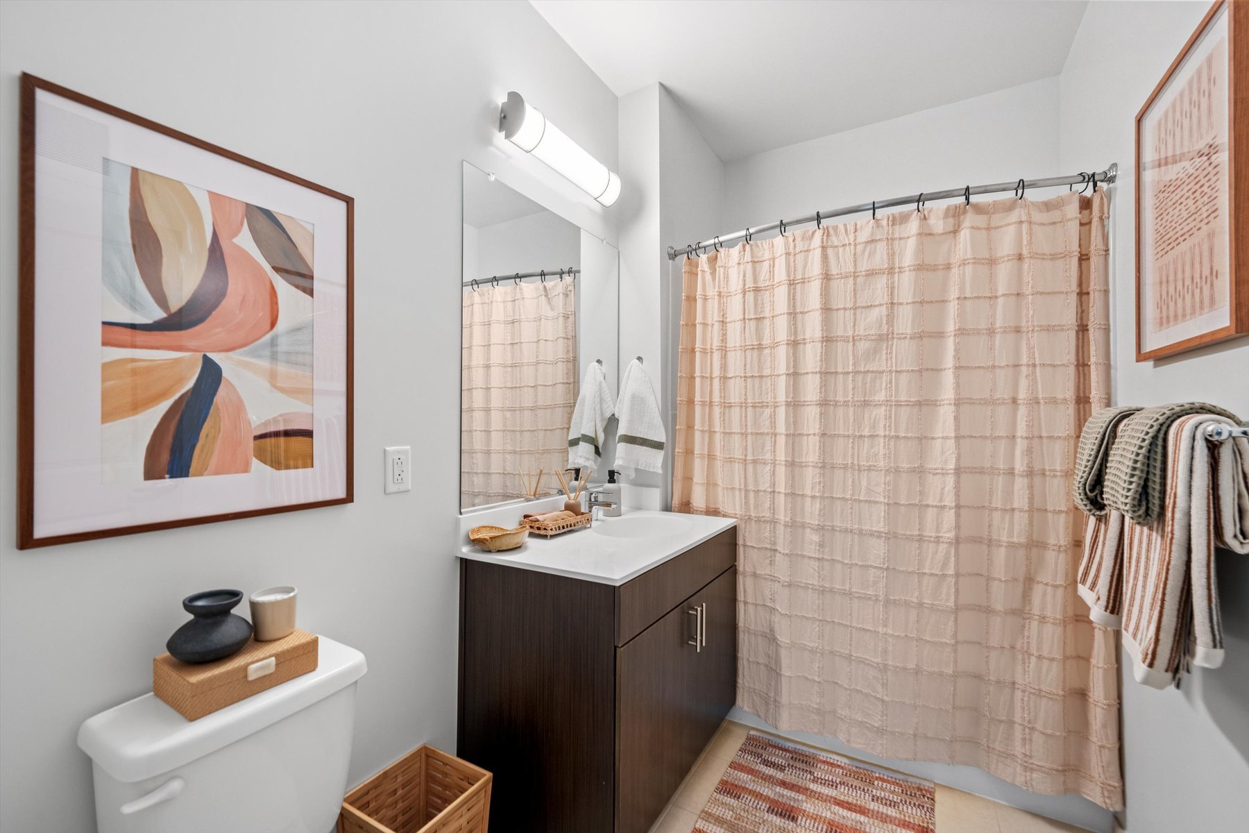 Modern bathroom at Landmark College Park with a dark wood vanity, white sink, large mirror, and overhead light. Abstract art hangs above the toilet. The shower has a peach curtain, and towels hang on wall hooks in this warm, neutral space.