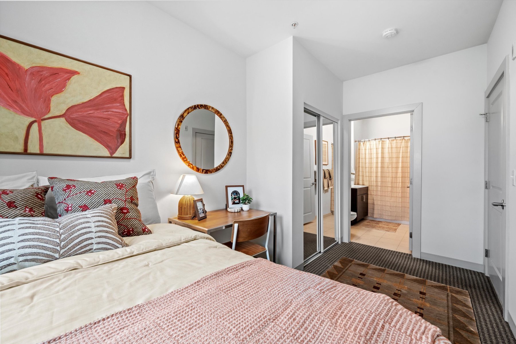 A cozy bedroom at Landmark College Park features a neatly made bed, patterned pillows, a wooden desk with a lamp and framed photo, a round mirror, and a large painting above the bed; a doorway leads to a bright bathroom.