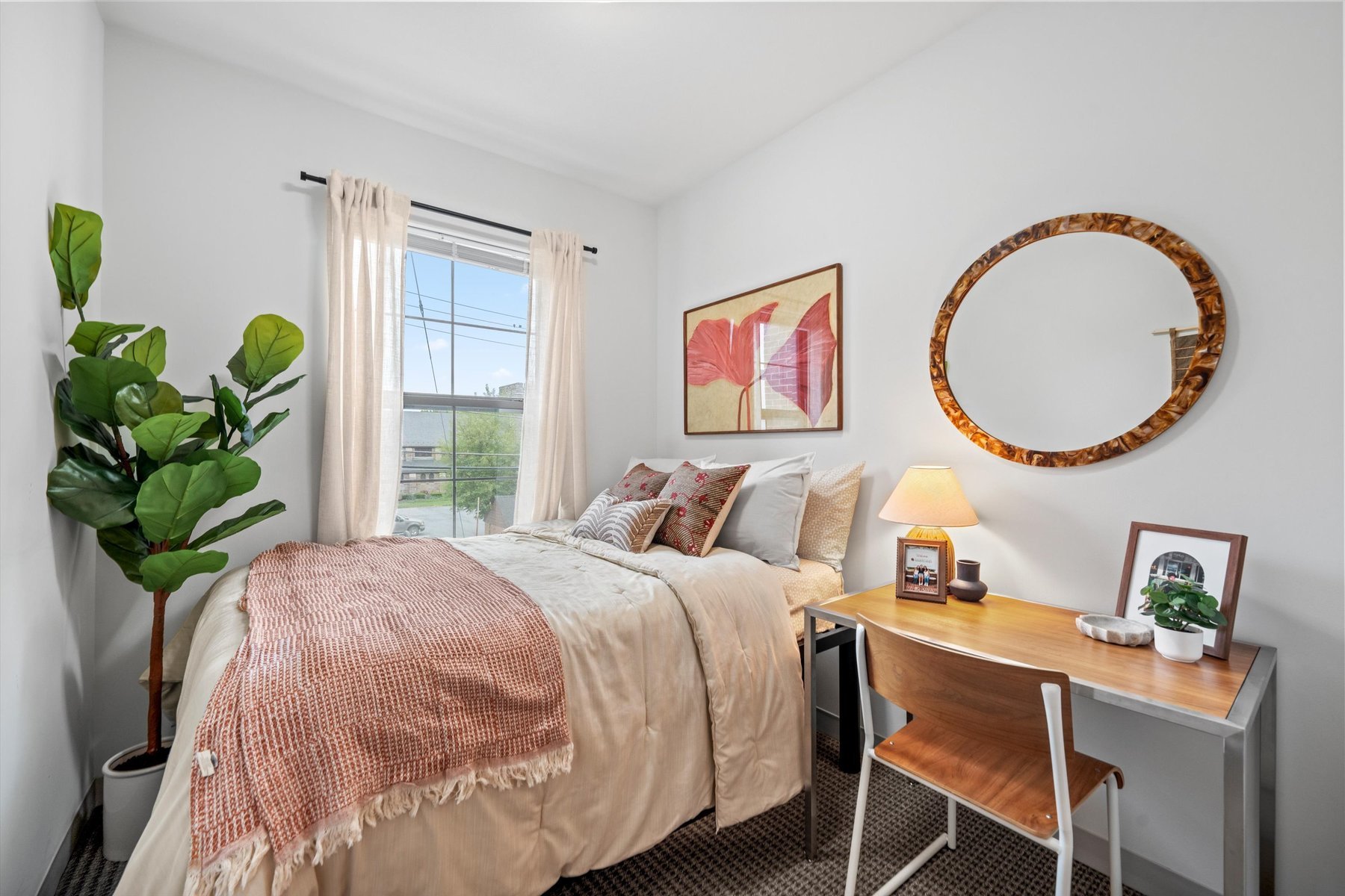 A cozy bedroom at Landmark College Park features a neatly made bed, a large green plant, a window with sheer curtains, a desk with a lamp and photo frame, a wooden chair, and wall art with red leaves and a round mirror.