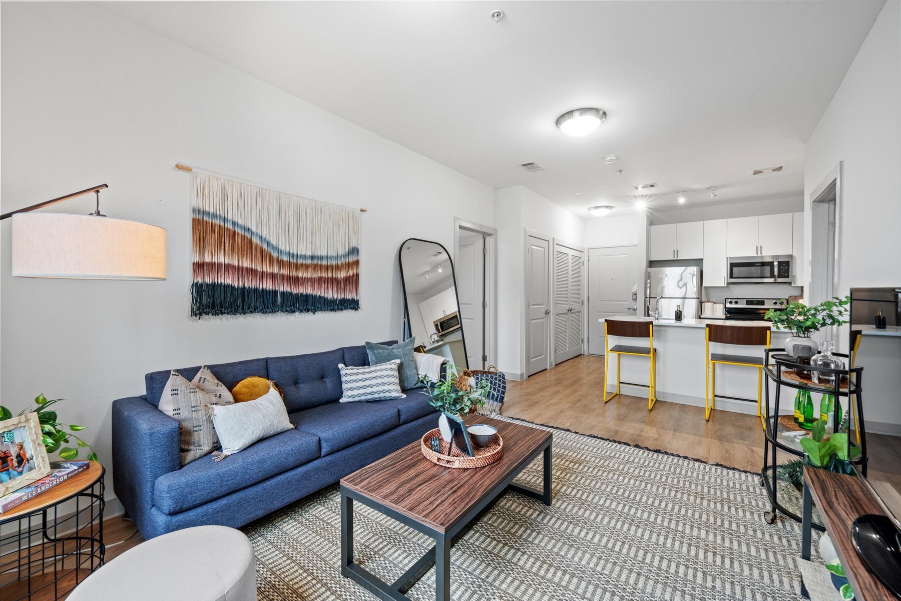 Modern open-concept living room and kitchen at Landmark College Park with a blue sofa, round mirror, and cozy decor. The kitchen features white cabinets, stainless steel appliances, and two yellow barstools at the island.