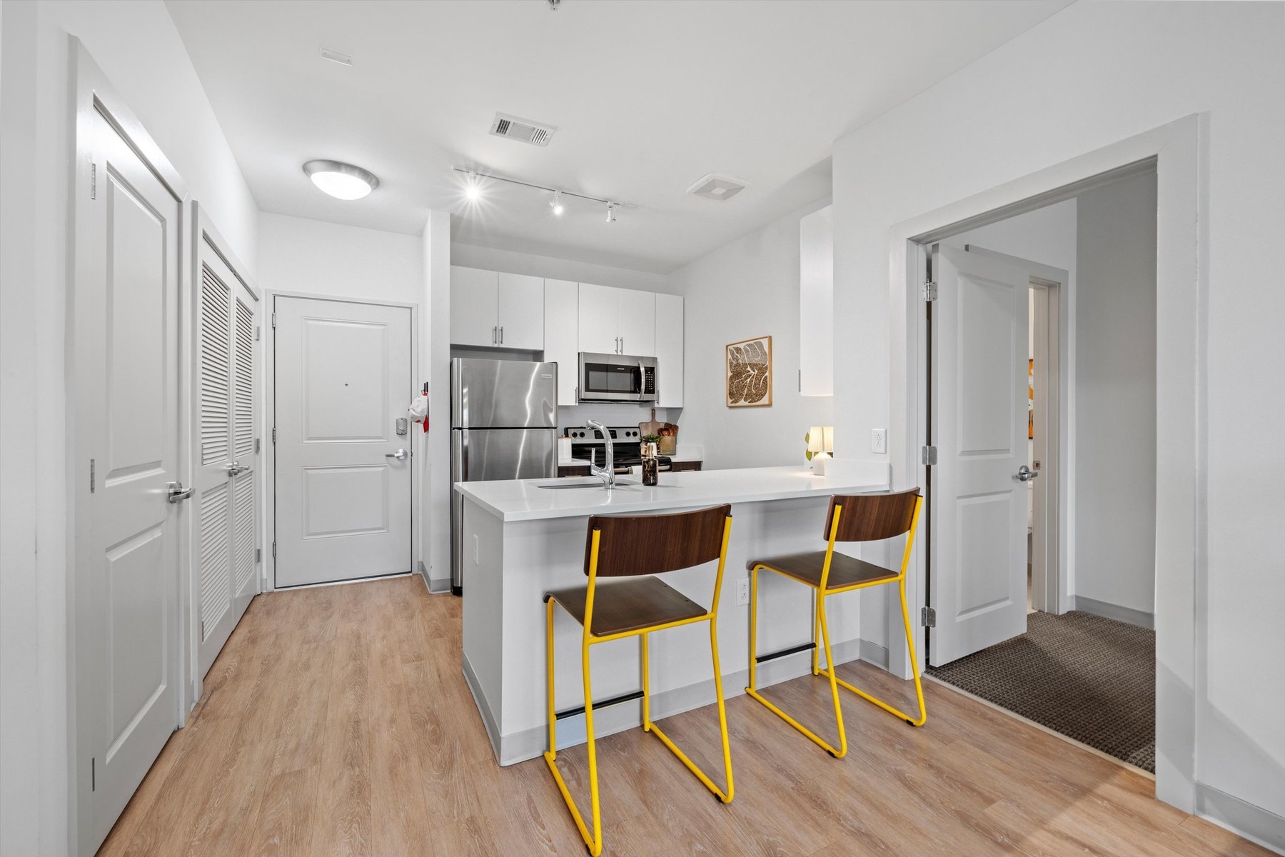Modern kitchen at Landmark College Park with white cabinets, stainless steel appliances, an island with yellow bar stools, light wood flooring, and open doors to adjacent rooms. Bright, clean, and minimalistic design.