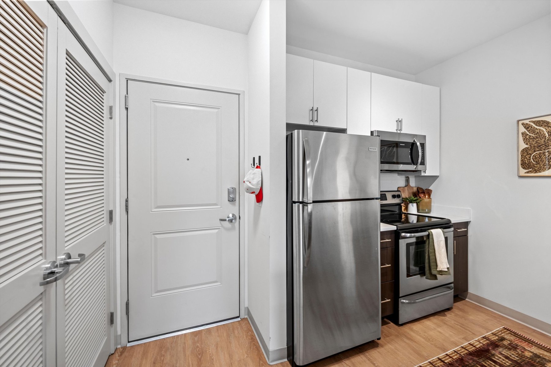 Modern kitchen at Landmark College Park with stainless steel refrigerator, oven, and microwave, white cabinets, wooden floor, and a closed white entry door with hooks holding hats next to the kitchen area.