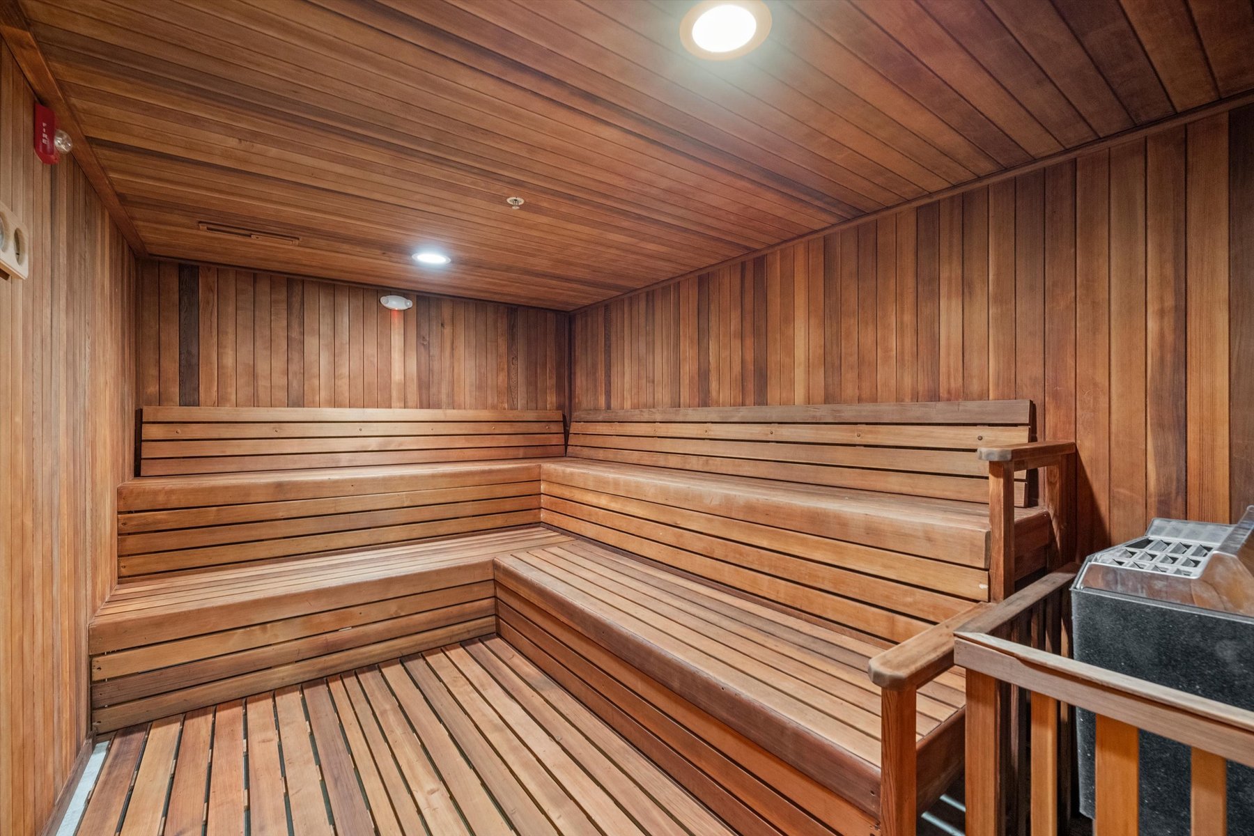Spacious wooden sauna at Landmark College Park with tiered bench seating, warm lighting from ceiling fixtures, and a heater in the corner, all surrounded by smooth wood-paneled walls and flooring.