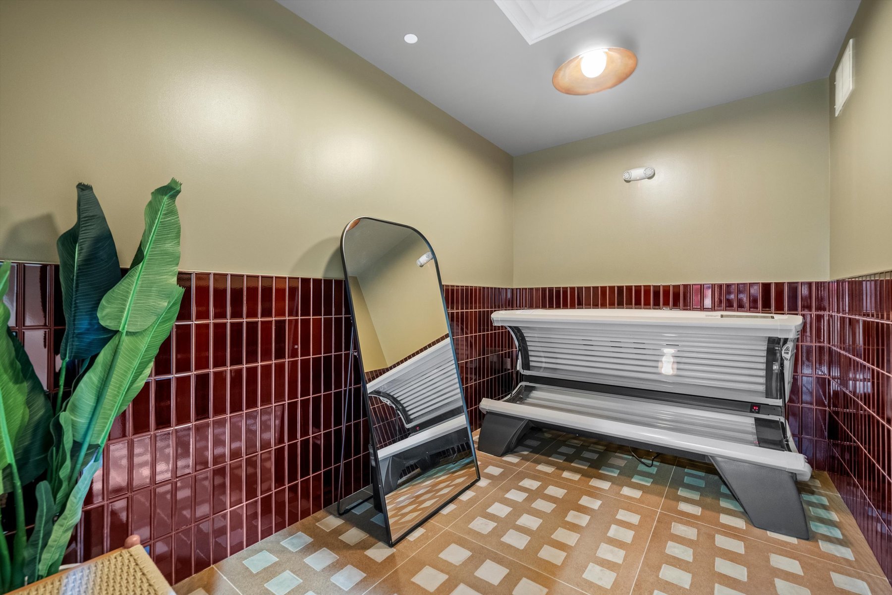 A room at Landmark College Park features red tile walls, a tanning bed, a large standing mirror leaning against the wall, a potted plant, and patterned floor tiles under soft ceiling lighting.
