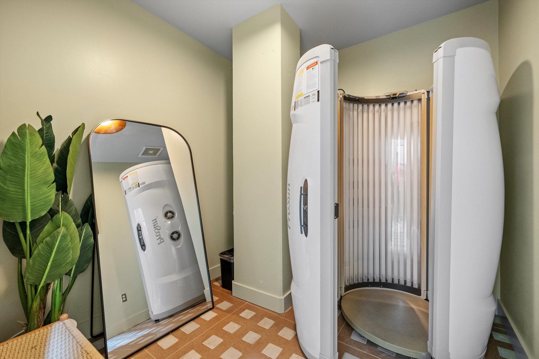 A room at Landmark College Park with a vertical tanning booth, a large leaning mirror, a green plant, and tiled flooring. The booth is open, revealing its interior lights and controls. The walls are painted a soft green.