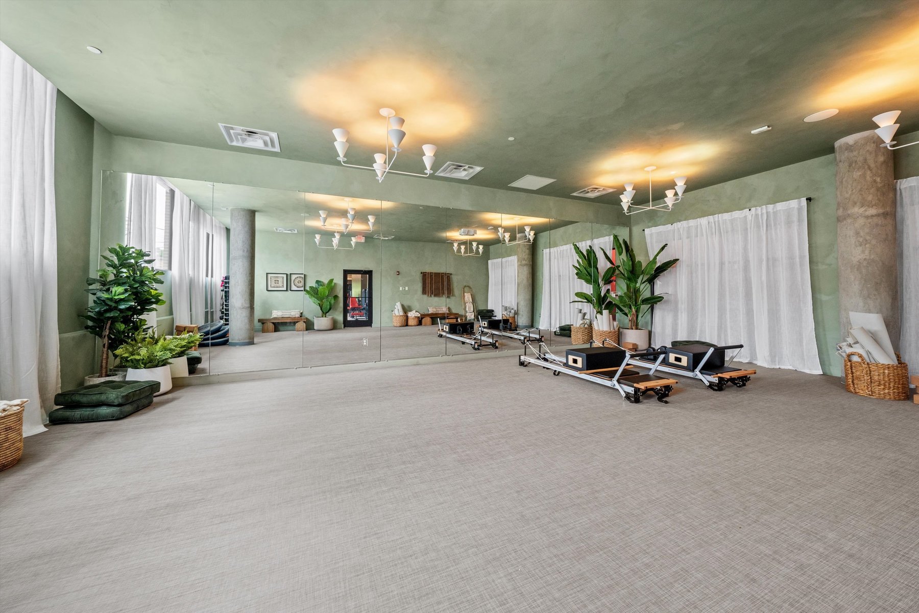 Spacious fitness studio at Landmark College Park with mirrored wall, soft green walls and ceiling, two pilates reformer machines, potted plants, woven baskets, and white curtains creating a calm, serene atmosphere.