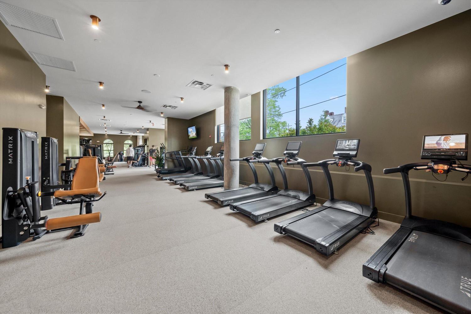 A modern gym at Landmark College Park features treadmills lined up by windows, strength training machines on the left, TVs mounted on the walls, and natural light streaming in from large windows.