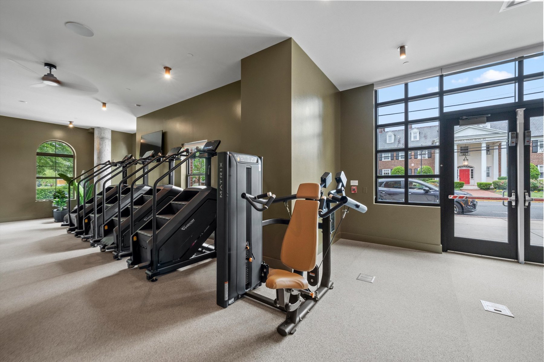 A modern gym at Landmark College Park with brown walls features several treadmills and a seated exercise machine near large windows and a glass door overlooking buildings and parked cars outside.