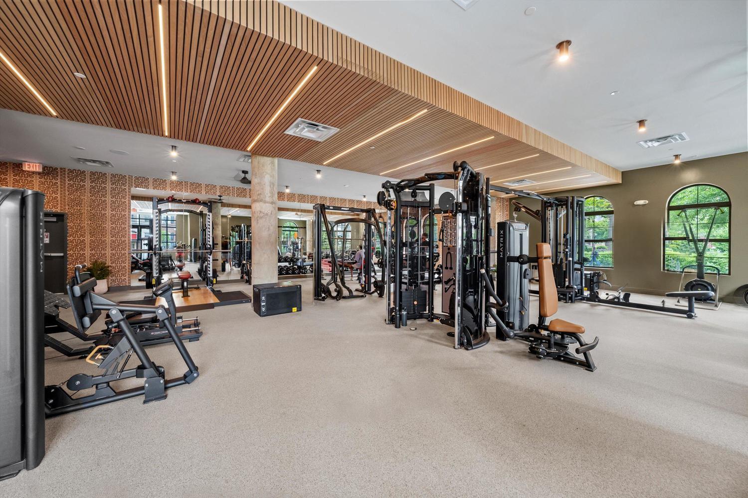 Spacious gym at Landmark College Park with exercise machines, weight equipment, mirrors, and large windows letting in natural light. The room features modern decor and stylish wood-paneled ceiling accents.
