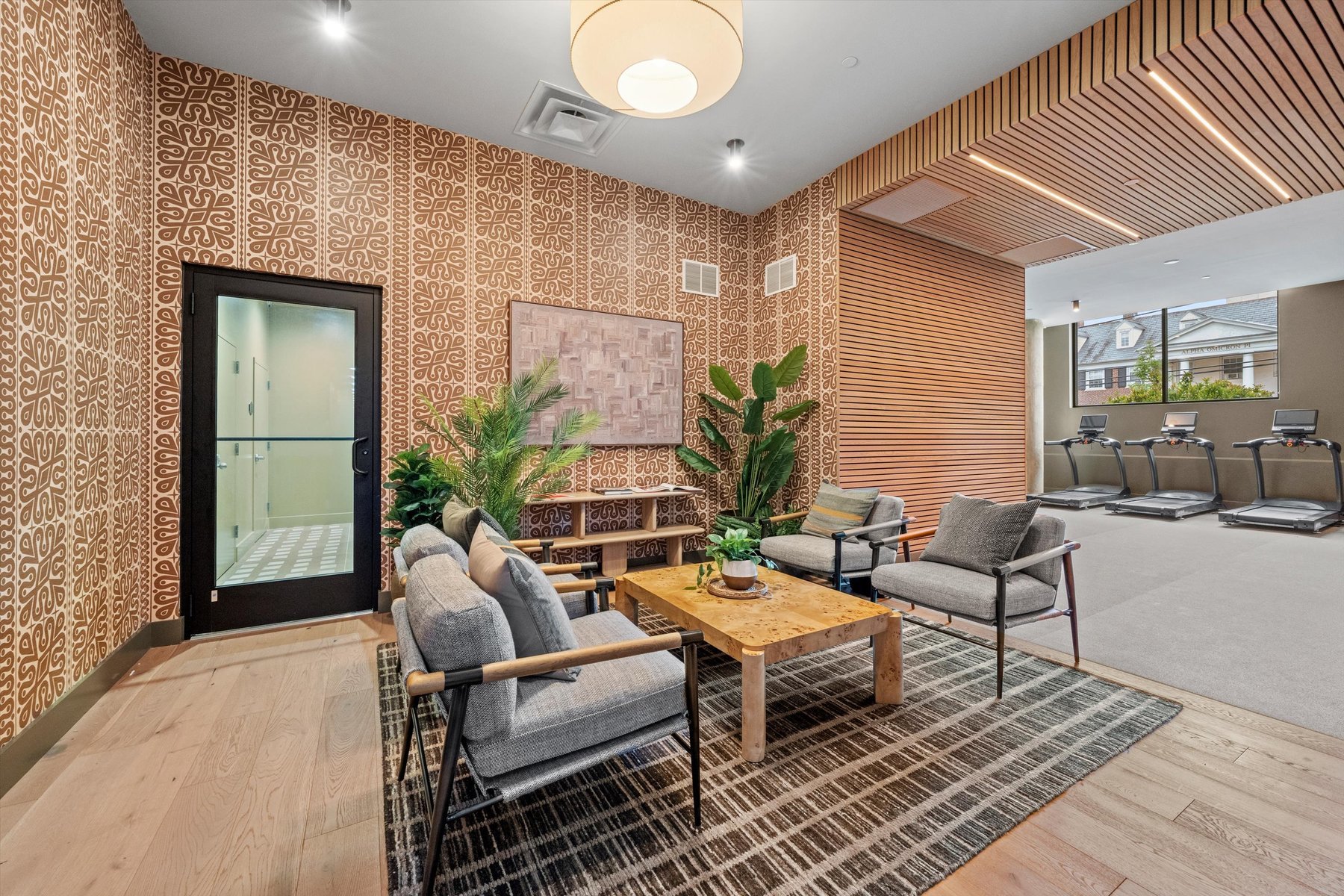 Modern lounge area with patterned walls, gray armchairs, a wooden coffee table, potted plants, and a rug at Landmark College Park; adjacent to a fitness room with treadmills visible through an open wooden slat partition.