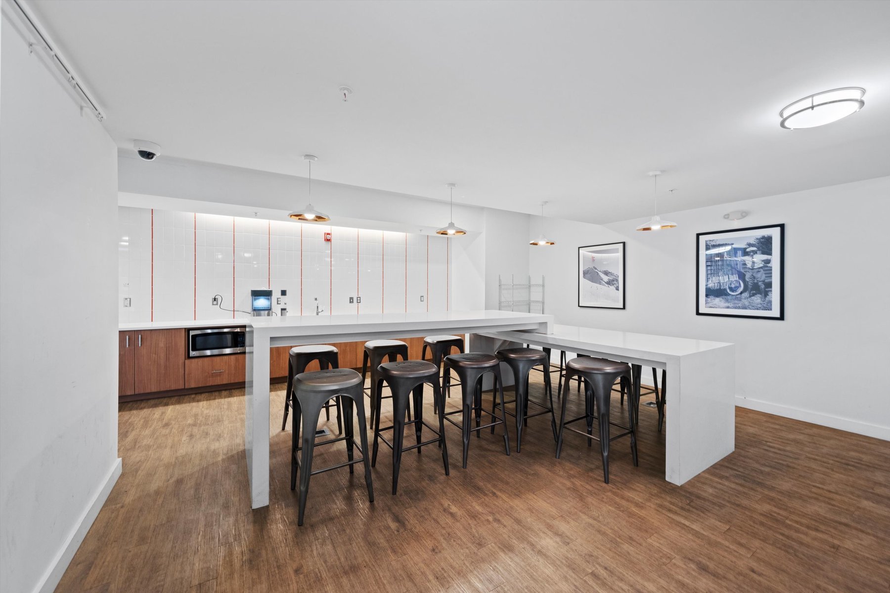 Modern break room at Landmark College Park with white walls, a long white table, black metal stools, wooden floors, and a kitchenette with microwave. Pendant lights illuminate the space as two large framed photographs decorate the wall.