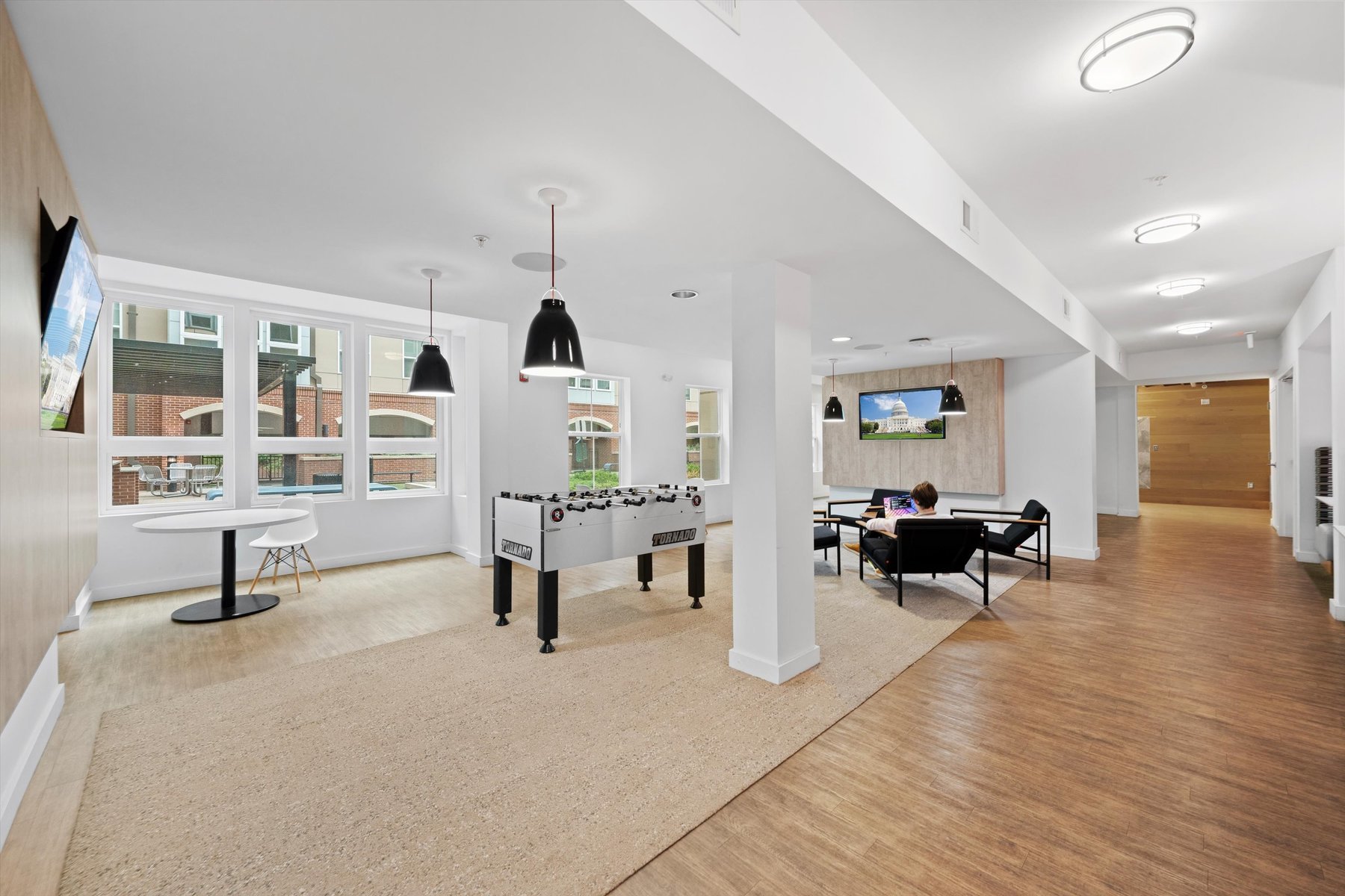 Modern lounge at Landmark College Park with a foosball table, round table and chairs by large windows, and people relaxing on a sofa near a TV. The space features wood-style flooring, white walls, and stylish pendant lights.