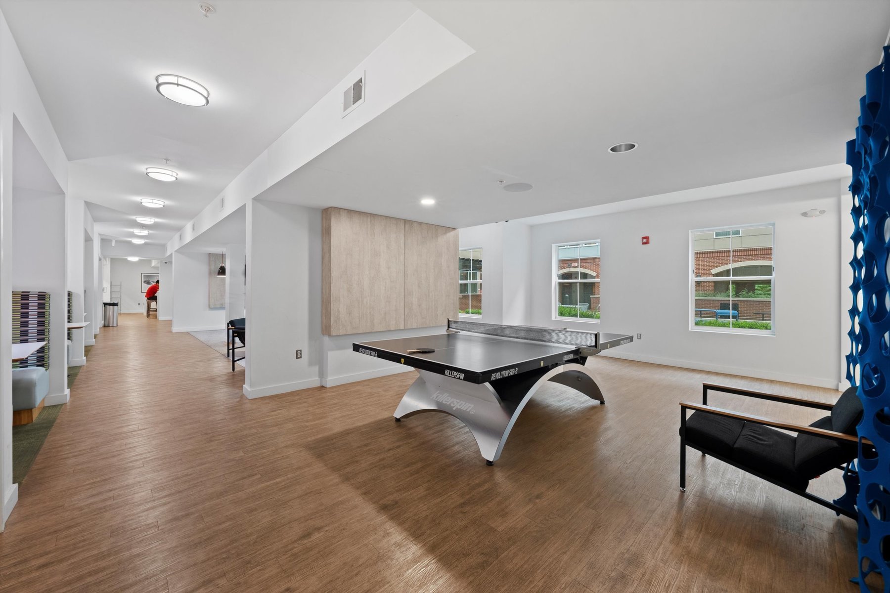 A modern recreation room at Landmark College Park features wood flooring, a black ping pong table in the center, large windows streaming natural light, a black chair, and a hallway visible to the left.