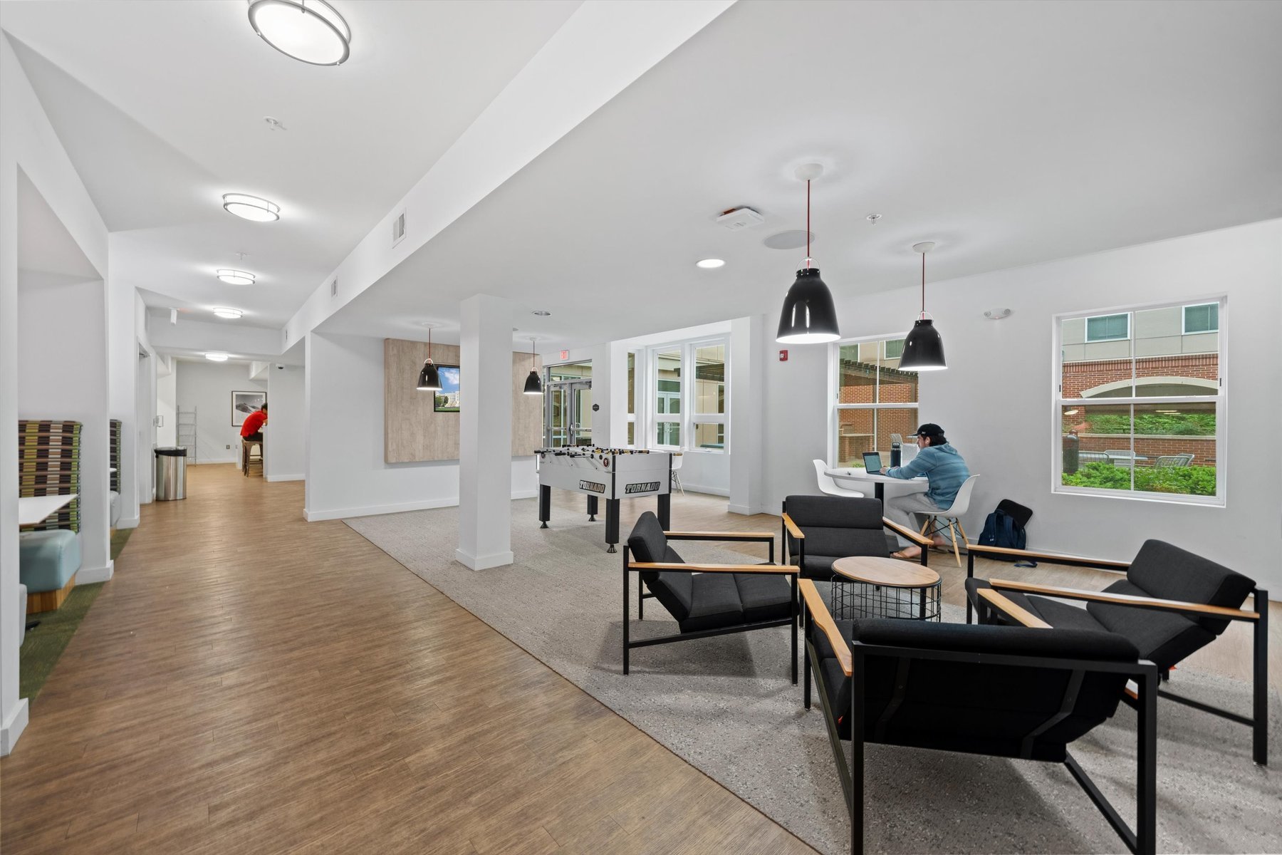Modern common area at Landmark College Park with black armchairs, tables, pendant lights, and large windows; one person working on a laptop at a table, another walking near the foosball table in the background.