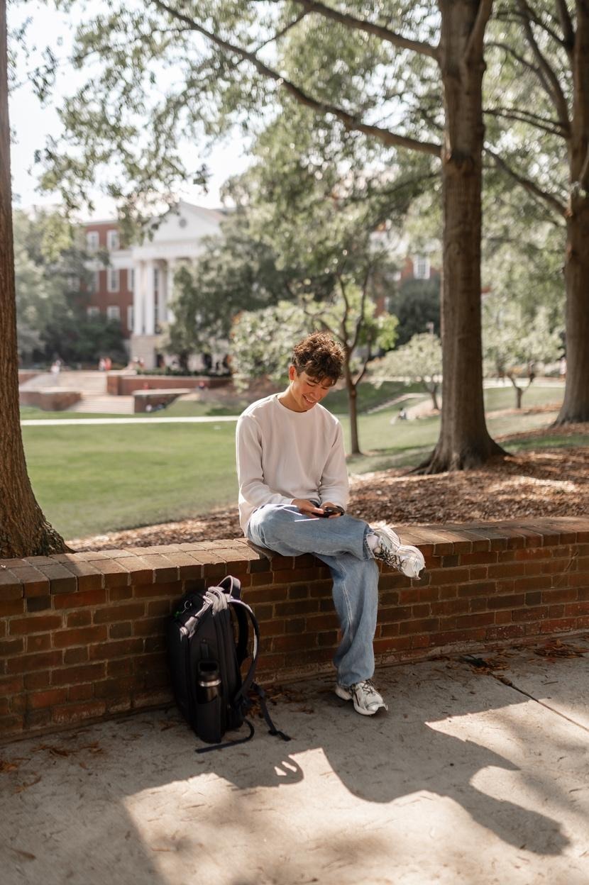 A young man in a white shirt and jeans sits on a brick ledge outdoors, looking at his phone near student apartments near campus. His backpack rests nearby, with trees and a grassy campus area in the background.